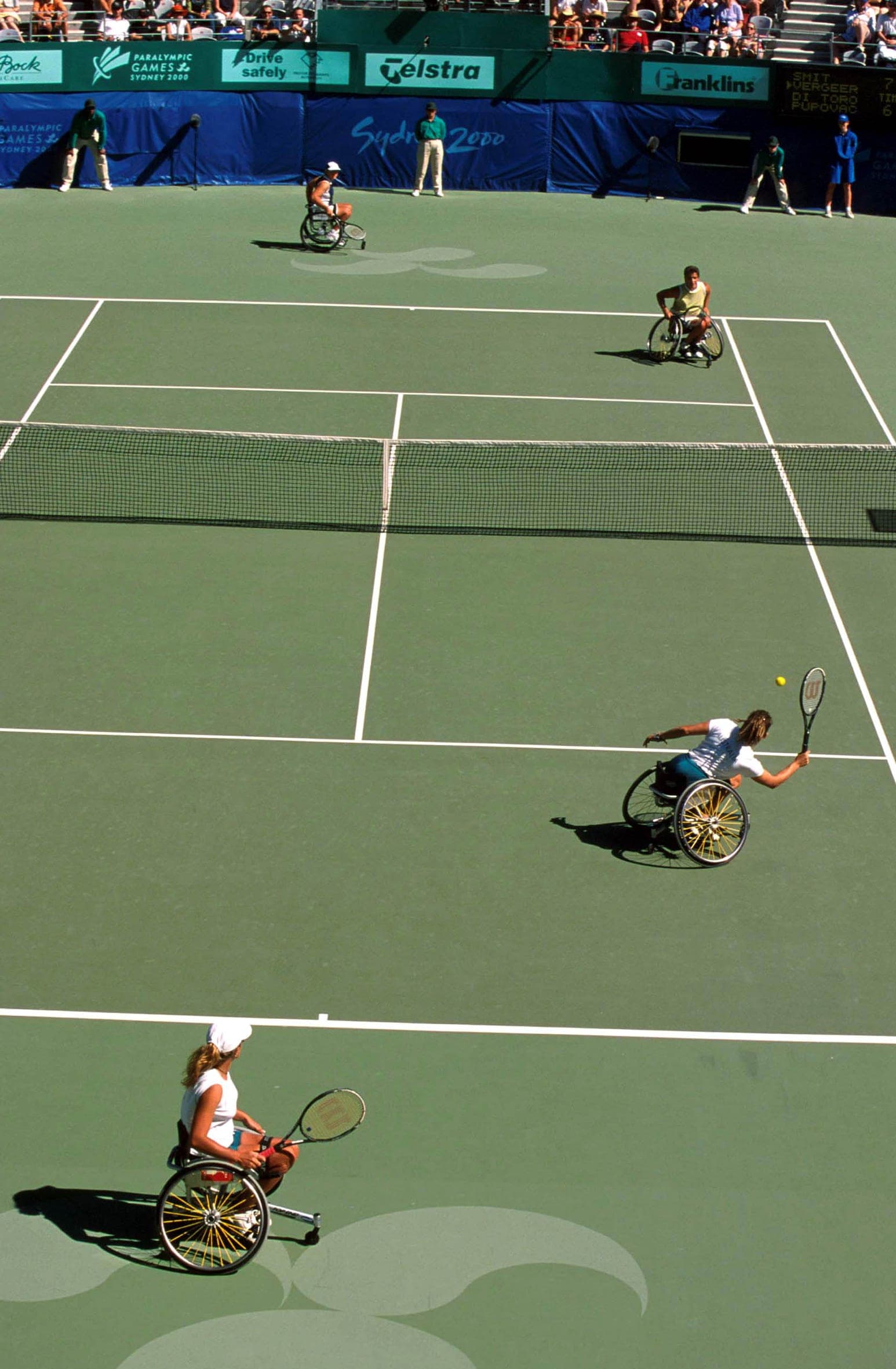 A wheelchair tennis player reaches for the ball during a match, seen from above the court.