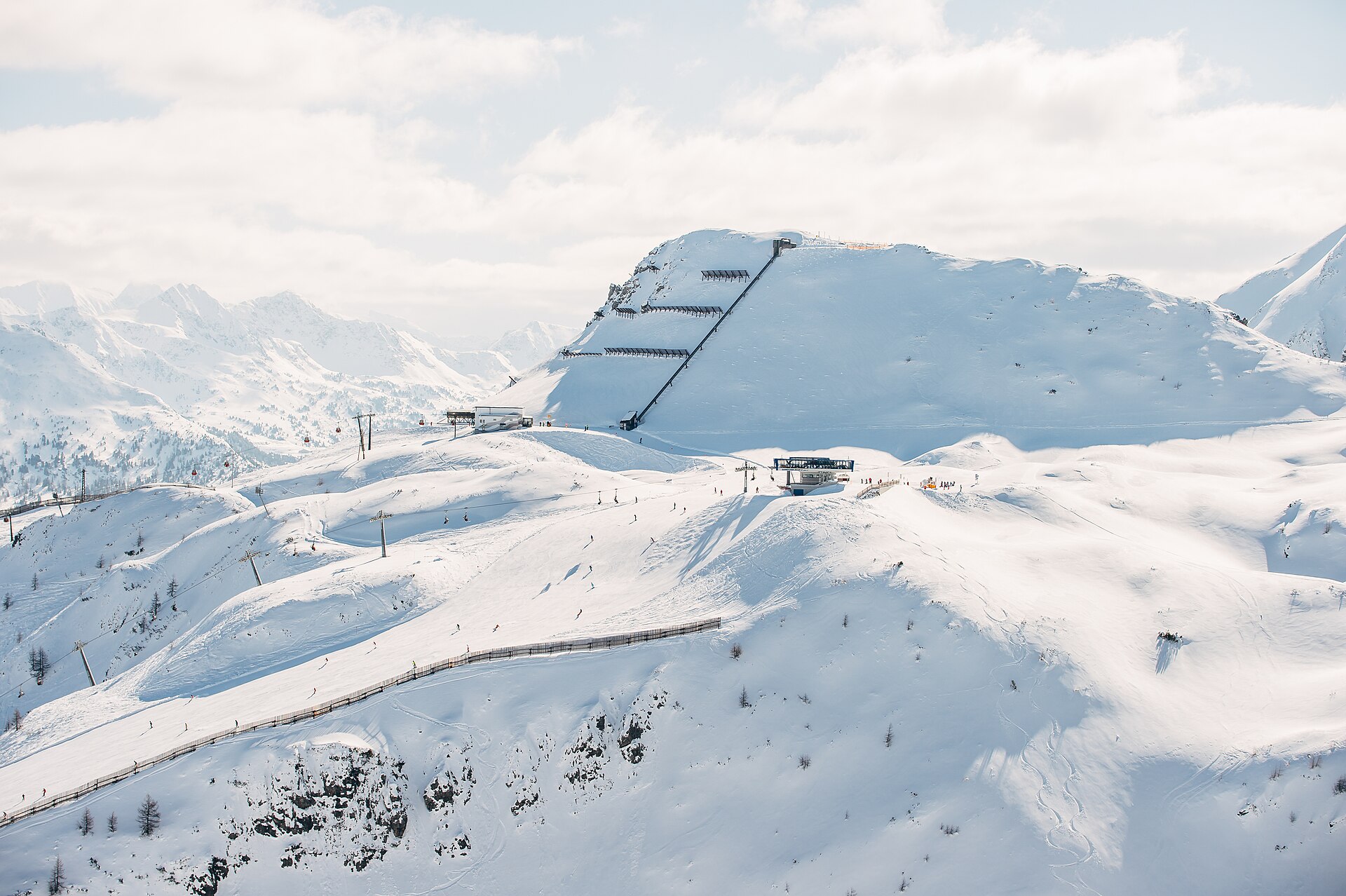 A snow-covered Alpine ski area seen from above: groomed pistes crossed by chairlifts and cable cars, a line of avalanche fencing along a ridge, and a steep terraced face rising to a peak beyond.