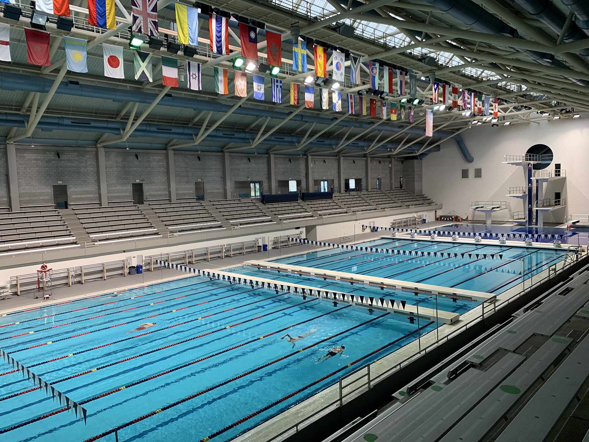 An indoor competition swimming pool with lane ropes in place, spectator seating alongside and a diving well beyond.