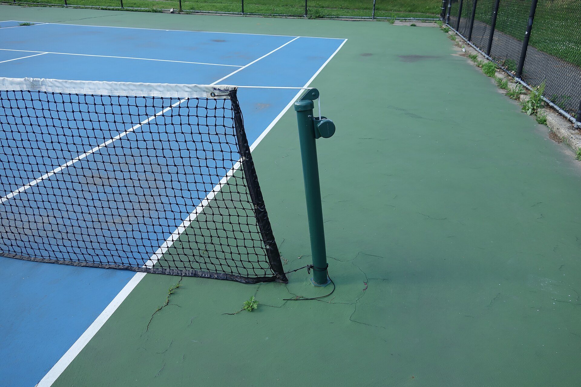The end of a tennis net at its post on a blue and green hard court: a round green post set outside the sideline, the dark netting with a white band along its top, and the doubles sideline running underneath.
