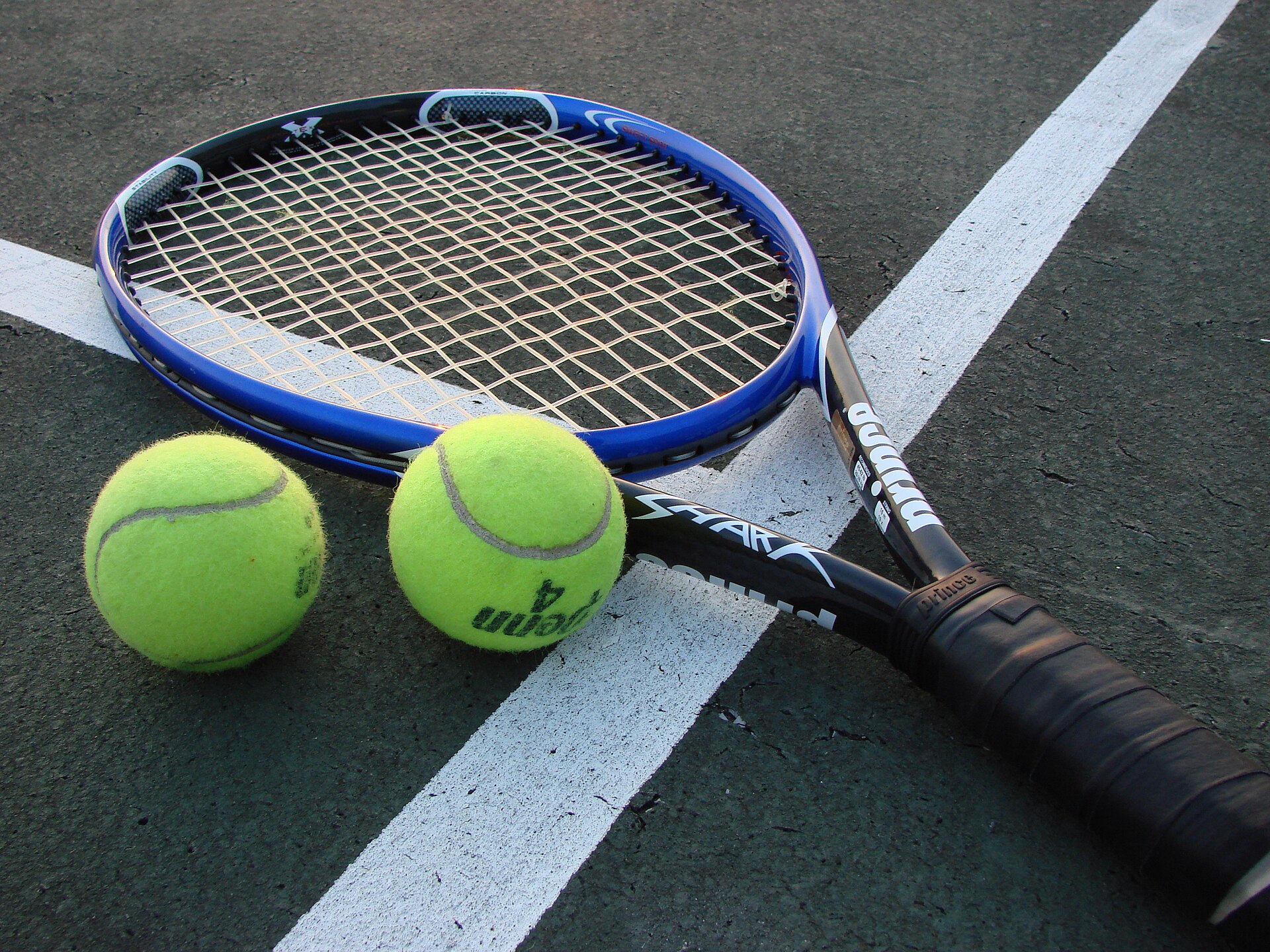 A tennis racket lying on a court surface next to two tennis balls.